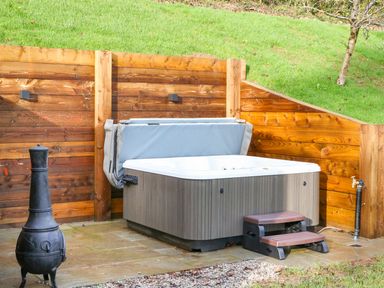 An outdoor hot tub with cover and steps on a stone patio next to a metal chiminea and wooden fence with grass background at Little Silver Fox in High Bickington