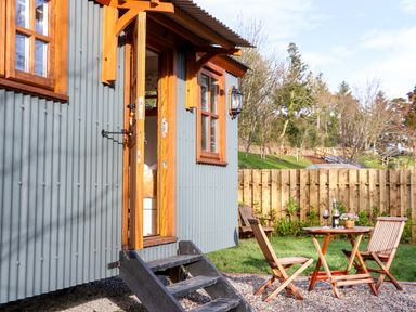 An outdoor area with wooden chairs and a table with wine glasses next to a grey corrugated metal building with wooden framed windows and door at Little Silver Fox in High Bickington