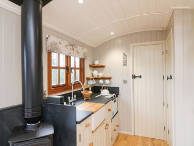 A small kitchen with a wood stove sink stove wooden shelves and a window with animal print curtain at Little Silver Fox in High Bickington