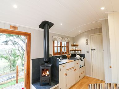 A small kitchen with a wood burning stove a window shelves with cups and teapots and a closed door at Little Silver Fox in High Bickington