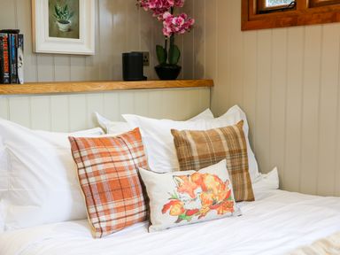 A bed with white sheets and three decorative cushions under a wooden shelf with books and a vase of flowers at Little Silver Fox in High Bickington