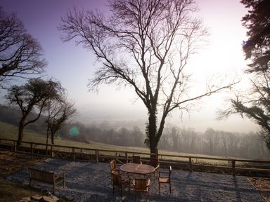 An outdoor seating area with a round wooden table and six chairs on gravel surrounded by trees overlooking a foggy field