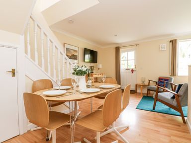 A dining area with a wooden table and six chairs set with plates and glasses next to a sitting area with two armchairs on a blue rug at Pugwash Cottage in Falmouth
