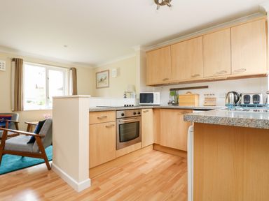 A kitchen with wooden cabinets a built in oven microwave kettle and toasters next to a sitting area with armchairs at Pugwash Cottage in Falmouth