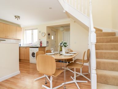 A dining area with a wooden table set for four and a kitchen with cupboards and a washing machine near stairs at Pugwash Cottage in Falmouth