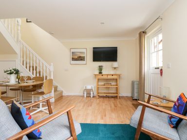 A living room with two armchairs a wooden table under a wall-mounted TV and a dining table near a staircase at Pugwash Cottage in Falmouth