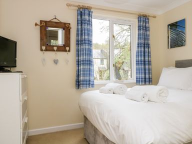 A bedroom with a bed with white linens and rolled towels near a window with blue plaid curtains and a dresser with a television at Pugwash Cottage in Falmouth