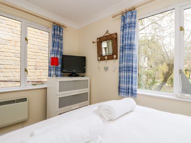 A bedroom with a bed with rolled white towels a cabinet with a television blue checked curtains and a window with a garden view at Pugwash Cottage in Falmouth