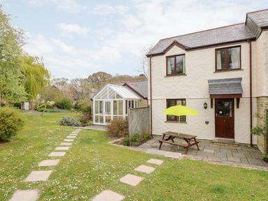 An exterior view of a two-story house with a wooden door a picnic bench with a yellow umbrella on a paved patio and a garden path leading to a glass conservatory at Pugwash Cottage in Falmouth
