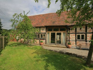 A garden with trees and grass outside a brick and timber building with wooden doors and windows at Whites Farm Barn in Ledbury