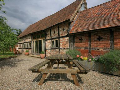 An outdoor area with a wooden picnic table on a gravel surface next to a brick and timber building with flower beds at Whites Farm Barn in Ledbury