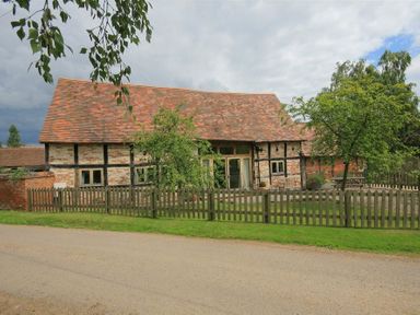 A brick and timber house with a tiled roof behind a wooden fence and trees at Whites Farm Barn in Ledbury