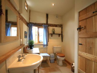 A bathroom with a sink toilet a chair table blue curtains and a wooden door at Whites Farm Barn in Ledbury
