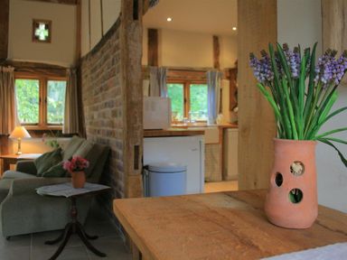 A sitting area with a green sofa and a small round table with flowers near a brick wall and a kitchen in the background at Whites Farm Barn in Ledbury