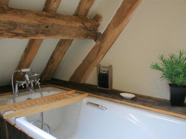 A bathtub with a wooden bath tray, faucet, bottles, a dish, and a potted plant under wooden ceiling beams at Whites Farm Barn in Ledbury