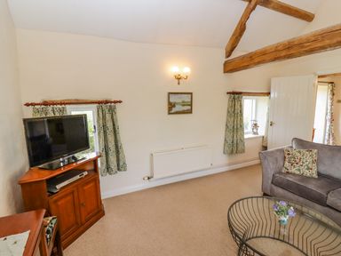 A living room with a grey sofa and floral cushions a glass coffee table a TV on a wooden stand and wooden ceiling beams at Pheasant Cottage in Minster Lovell