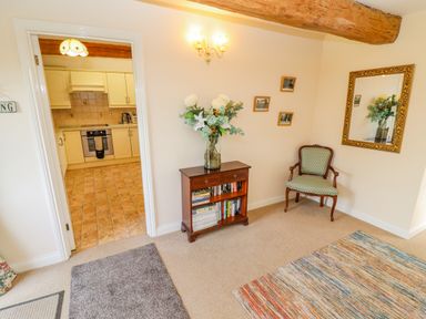 A room with a wooden bookshelf holding books and a vase of flowers next to a green upholstered chair and a mirror with a gold frame in Pheasant Cottage Minster Lovell