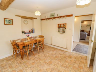 A dining room with a wooden table and chairs a wall shelf with cups and towels at Pheasant Cottage in Minster Lovell