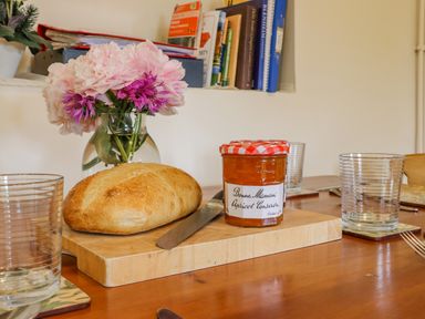 A wooden dining table with a loaf of bread, a jar of apricot conserve, a knife, glass cups, and a vase of flowers at Pheasant Cottage in Minster Lovell