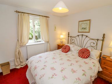 A bedroom with a double bed floral bedding two red circular pillows wooden bedside tables with lamps and a window with curtains at Pheasant Cottage in Minster Lovell