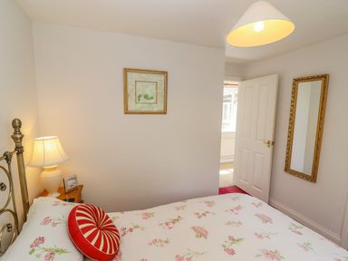 A bedroom with a bed with floral bedding a bedside table with lamp and picture frame a wall mirror and an open door at Pheasant Cottage in Minster Lovell