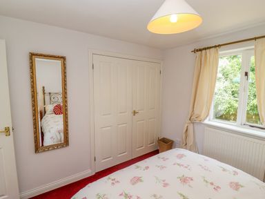 A bedroom with a floral bedspread a full length mirror on the wall a window with curtains and a double door closet at Pheasant Cottage in Minster Lovell