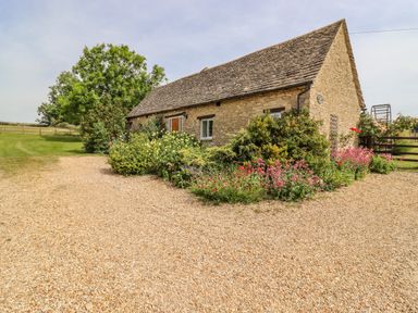 A stone cottage with flower beds and gravel driveway surrounded by greenery at Pheasant Cottage in Minster Lovell