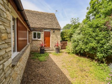 An exterior view of a stone cottage with a wooden door two potted plants a gravel path and green bushes at Pheasant Cottage in Minster Lovell