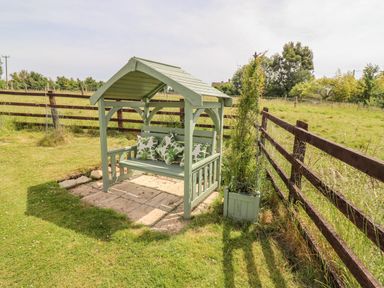 A green wooden garden bench with a roof and cushions next to a fence in a grassy yard at Pheasant Cottage in Minster Lovell