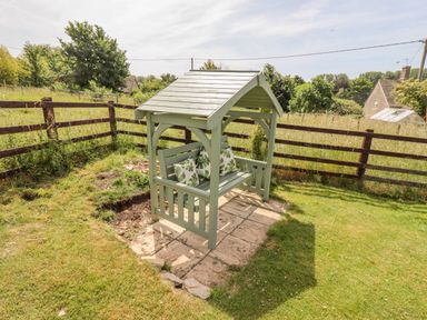 A wooden garden bench with a roof and cushions on a stone patio in a fenced grassy yard at Pheasant Cottage in Minster Lovell
