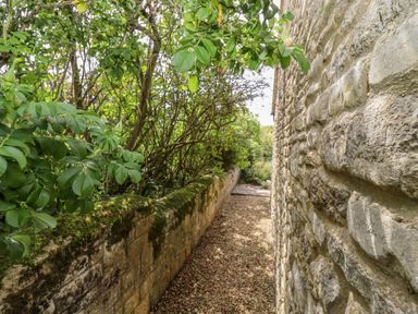 A narrow garden path with gravel between a stone wall and green foliage at Pheasant Cottage in Minster Lovell