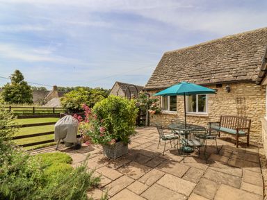 An outdoor patio with a table, chairs, umbrella, a bench, plants, and a covered barbecue grill at Pheasant Cottage in Minster Lovell