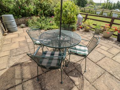 A patio with a round metal table and four chairs with cushions near plants and a wooden fence at Pheasant Cottage in Minster Lovell