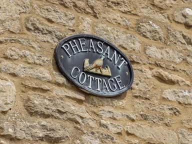 A stone wall with an oval sign that reads Pheasant Cottage with a pheasant image at Pheasant Cottage in Minster Lovell