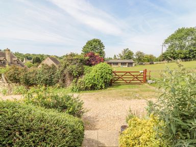 A garden with various bushes and a gravel path leading to a wooden gate and open fields at Pheasant Cottage in Minster Lovell