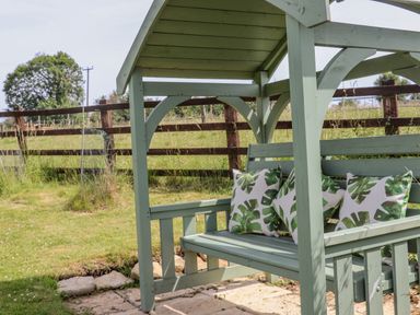 A green wooden garden swing with leaf patterned cushions in a grassy yard with a wooden fence in the background at Pheasant Cottage in Minster Lovell