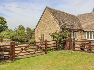 A stone cottage with a wooden fence and gate surrounding a garden with flowers at Pheasant Cottage in Minster Lovell