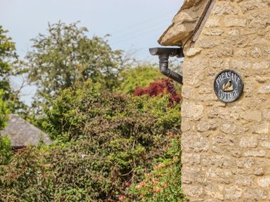 Exterior stone wall of a building with a sign that reads Pheasant Cottage surrounded by green shrubbery and trees at Pheasant Cottage in Minster Lovell