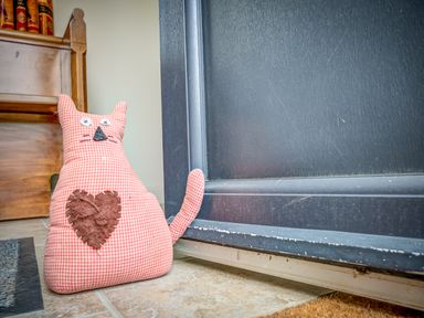 A fabric cat-shaped doorstop with a heart shape on tiled floor next to a black door and wooden furniture at Top Barn in Chipping Norton