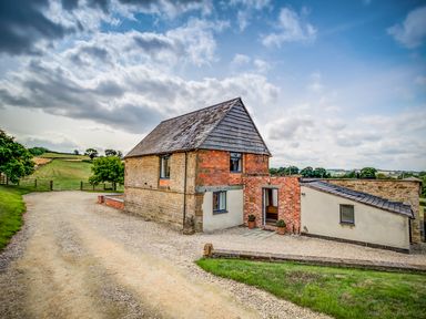 A barn-style building with brick and stone walls on a gravel driveway in a rural landscape at Top Barn in Chipping Norton