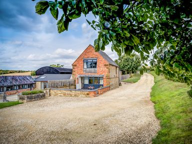 A brick and stone house with a gravel driveway and greenery at Top Barn in Chipping Norton