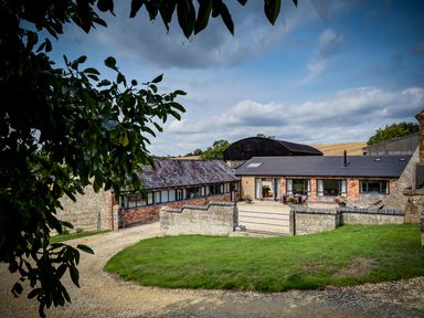 A courtyard with stone walls and a gravel driveway around a grassy area with farm buildings in the background at Top Barn in Chipping Norton