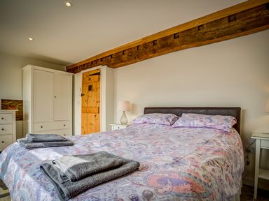 A bedroom with a double bed with patterned bedding two bedside tables with lamps a wooden door a white wardrobe and exposed wooden beam at Top Barn in Chipping Norton