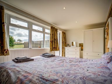 A bedroom with a patterned bedspread two folded towels on the bed white dresser and wardrobe near windows with curtains at Top Barn in Chipping Norton