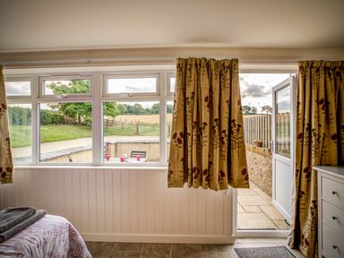 A room with a bed, floral curtains, window, door opening to a patio with chairs and a table at Top Barn in Chipping Norton