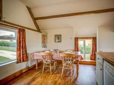 A dining area with a wooden table and four chairs next to windows with curtains at Top Barn in Chipping Norton