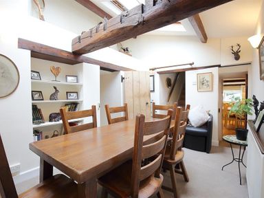 A dining room with a wooden table and six chairs with exposed wooden beams and a built-in shelf with decor at Pike Cottage in Stow-On-The-Wold
