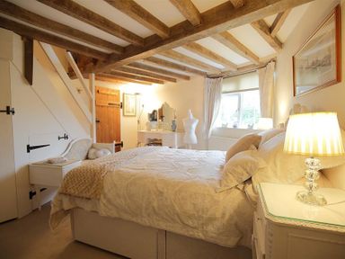 A bedroom with a bed bedside tables lamps a vanity table with a mirror a window and exposed wooden ceiling beams at Pike Cottage in Stow-On-The-Wold