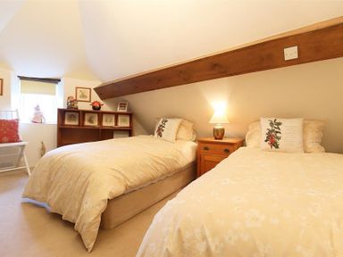 A bedroom with two single beds a wooden bedside table with a lamp a small window a wooden shelf and a chair at Pike Cottage in Stow-On-The-Wold