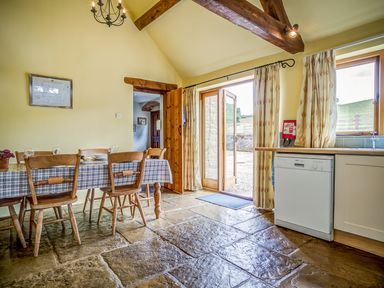 A kitchen and dining area with a checkered tablecloth on a wooden dining table and wooden chairs near a door and window at Little Barn in Ascott near Chipping Norton